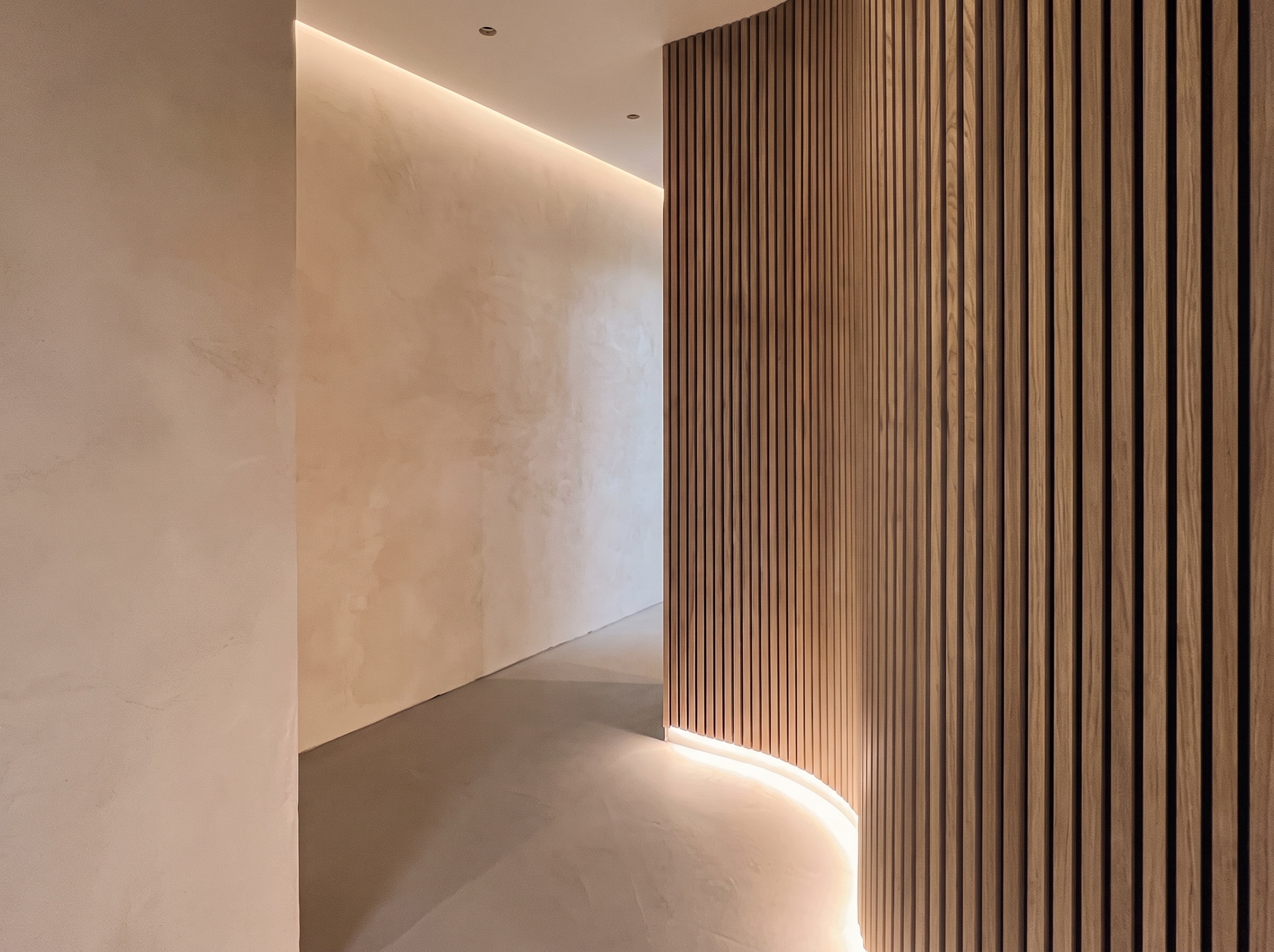 Curved wood-slat partition wall in a hallway with cove lighting, installed by Maynard